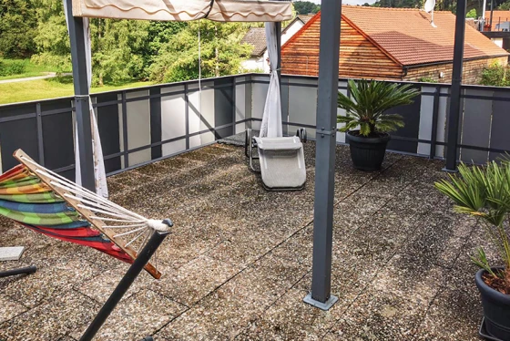 Die Dachterrasse mit alten Waschbetonplatten bietet eine großzügige Aufenthaltsfläche. Die verschmutzte Oberfläche der Waschbetonplatten zeigt deutliche Gebrauchsspuren. Der Sonnenschutz spendet an heißen Tagen angenehmen Schatten. Pflanzkübel mit Palmen und eine Hängematte schaffen eine einladende Atmosphäre. Die Geländerkonstruktion aus Metall und Glas rundet das moderne und offene Design der Terrasse ab.