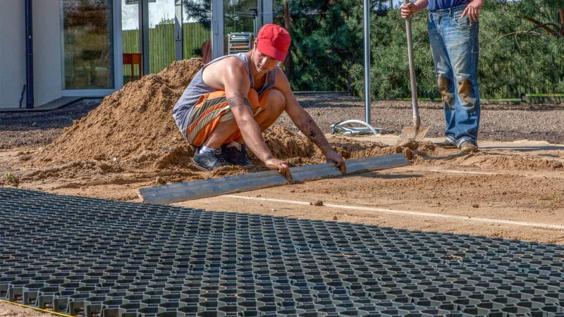 Dans le jardin, une couche de base de grilles en nid d’abeille en plastique est disposée sur un lit de sable. Un jeune homme nivelle le sable.