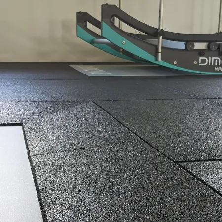 Das Bild zeigt einen modernen Fitnessbereich, der mit hochwertigen WARCO Fitnessbodenplatten ausgestattet ist. Der Bodenbelag besteht aus strapazierfähigem Gummi, das eine rutschfeste und stoßdämpfende Oberfläche bietet, ideal für intensive Trainingseinheiten. Der Boden ist in einem eleganten, mattschwarzen Finish gehalten, das sowohl funktional als auch ästhetisch ansprechend ist. Im Hintergrund des Bildes ist ein Trainingsgerät mit der Aufschrift "DIMOVE" zu sehen, das auf dem rutschfesten Bodenbelag sicher positioniert ist. Die klaren Linien der Bodenplatten und das minimalistische Design des Raumes unterstreichen die professionelle und hochwertige Ausführung des Fitnessbereichs, der sowohl für Einzel- als auch für Gruppentraining geeignet ist.