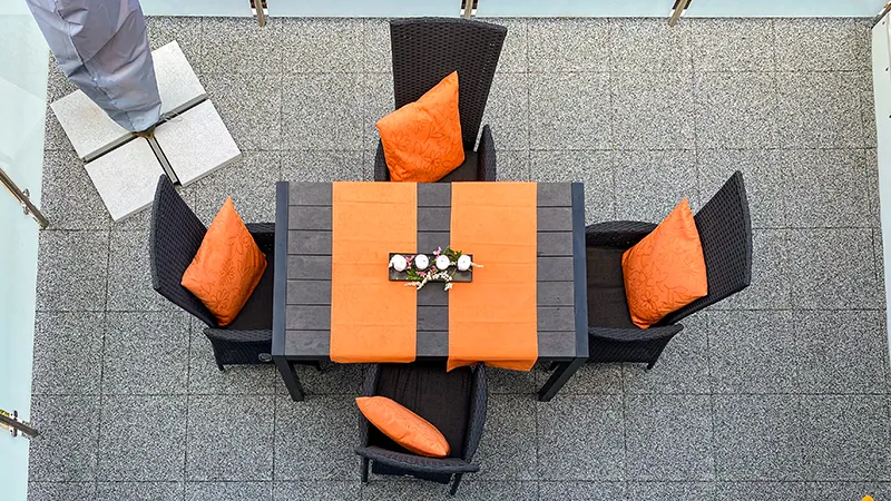 Op deze foto zie je een ruim balkon met grijze WARCO-vloerplaten. In het midden staat een grote eettafel, omringd door zwarte rotanstoelen met oranje kussens als kleuraccent. De elastische, antislip vloer zorgt voor comfort en een veilige ondergrond. Bovendien is de vloer onderhoudsvriendelijk en weerbestendig, wat de buitenruimte een moderne uitstraling geeft en uitnodigt om te ontspannen.
