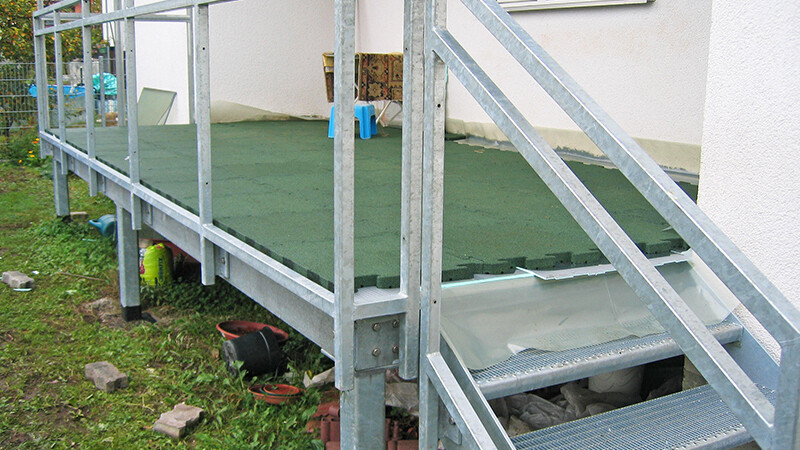 Metalen terras met groene platen grenzend aan een huis. Een kleine trap leidt twee treden omhoog. De onderbouw is gemaakt van traliewerk.