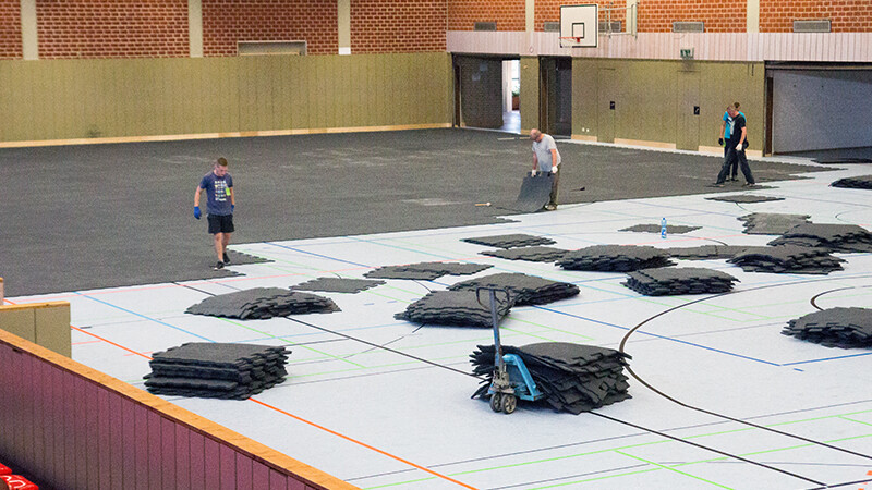 In einer großen Sporthalle wird ein Eventboden verlegt. Dieser besteht aus Gummigranulatmatten im Verbundpflaster-Design. Der Eventboden schützt den empfindlichen Sportboden der Halle vor Beschädigungen.