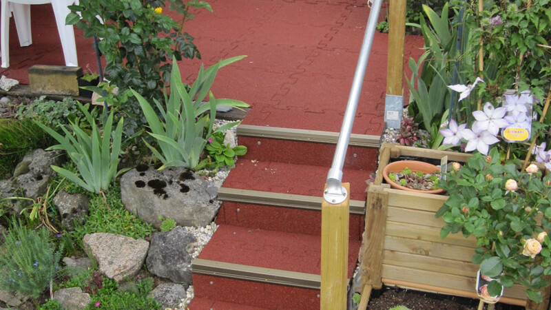 Een mooie rode trap leidt vanuit de tuin naar een terras. De rode Warco-platen passen goed bij de groene planten.