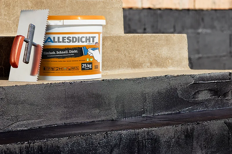 Das Bild zeigt einen 25-kg-Eimer der Gummiabdichtung ALLESDICHT von WARCO, der auf einer Betontreppe platziert ist. Der Eimer hat einen orangefarbenen Deckel und eine detaillierte Beschriftung, die das Logo von ALLESDICHT sowie den Slogan „Einfach. Schnell. Dicht.“ enthält, was auf die einfache, schnelle und dichte Abdichtung hinweist. Neben dem Eimer steht eine Zahnkelle, die für die manuelle Verarbeitung der Abdichtungsmasse verwendet wird. Die Treppenstufen sind teilweise bereits mit der schwarzen, elastischen Abdichtungsmasse beschichtet, wodurch eine wasserdichte und reißfeste Schutzschicht entsteht.