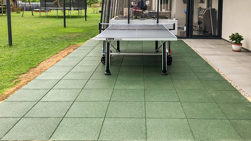 Sur une terrasse en dalles vertes WARCO, composées de granulats de caoutchouc liés au PU, se dresse une table de ping-pong devant une maison luxueuse. Dans le jardin, un trampoline et d’autres équipements invitent à la détente active et au mouvement. Ces dalles robustes et antidérapantes offrent confort et sécurité. Leur surface élastique préserve les articulations, tandis que leur structure facile d’entretien conserve un aspect soigné.