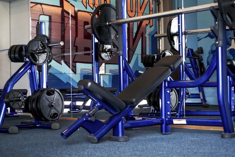 In een fitnessstudio kijkt men naar een blauw geschilderde hellingbank voor een spiegelwand. De schuine bank staat op een blauwe vloerbedekking. Dit zijn de vloerbeschermingsmatten van WARCO Fitness in het kleurendesign Atlantic.
