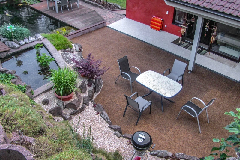 Een zitgedeelte bevindt zich op een terras dat bedekt is met aardkleurige vierkante terrastegels. Aan de rand van de foto zijn bijna natuurlijke vijvers en een begroeide oeverbescherming te zien.