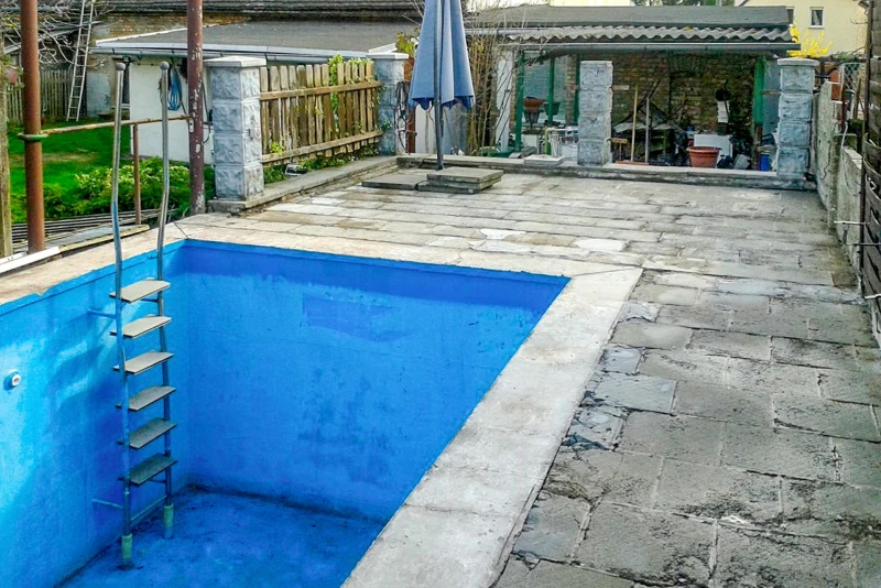 Een oud betonnen bassin in de grond met een betonnen rand wacht op renovatie
