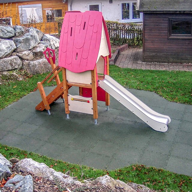 Auf einem Hanggrundstück wurde eine Ausbuchtung für einen kleinen Spielplatz, der aus einem auf grünen Fallschutzplatten aufgestellten Kleinkinder-Spielhaus mit Rutschbahn besteht, angelegt.