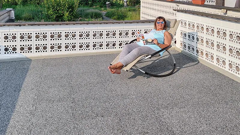 Op de foto is een ruim balkon te zien, dat is voorzien van grijze WARCO-vloerplaten. Iemand zit comfortabel in een schommelstoel, geniet van de zon en leest een boek. Het antislip en elastische oppervlak biedt zowel comfort als geluidsdemping. Bovendien is de vloer weerbestendig, robuust en onderhoudsvriendelijk, wat hem ideaal maakt voor langdurig gebruik. Zo ontstaat een uitnodigende buitenruimte om heerlijk te ontspannen.