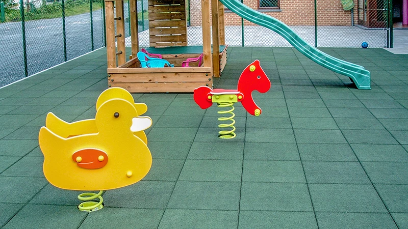 lik van een kinderspeelplaats waarvan het speeloppervlak volledig bedekt is met groene valbeveiligingstegels van WARCO. De kinderspeelplaats heeft verschillende speeltoestellen.