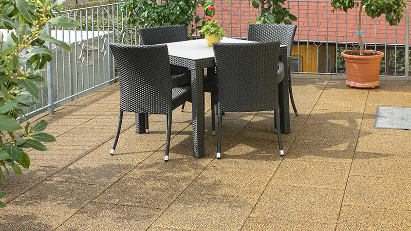 Sur une terrasse surélevée se trouve une table sombre entourée de quatre chaises tressées, mises en valeur par des dalles de terrasse WARCO d’un ton terracotta beige-brun. Ces dalles en granules de caoutchouc liées au PU offrent une surface antidérapante, amortissante et facile à entretenir. Des plantes en arrière-plan apportent une touche naturelle. Grâce à leur structure poreuse, le sol sèche rapidement après la pluie et garantit un confort de marche optimal.