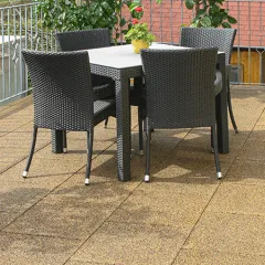 Sur une terrasse surélevée se trouve une table sombre entourée de quatre chaises tressées, mises en valeur par des dalles de terrasse WARCO d’un ton terracotta beige-brun. Ces dalles en granules de caoutchouc liées au PU offrent une surface antidérapante, amortissante et facile à entretenir. Des plantes en arrière-plan apportent une touche naturelle. Grâce à leur structure poreuse, le sol sèche rapidement après la pluie et garantit un confort de marche optimal.