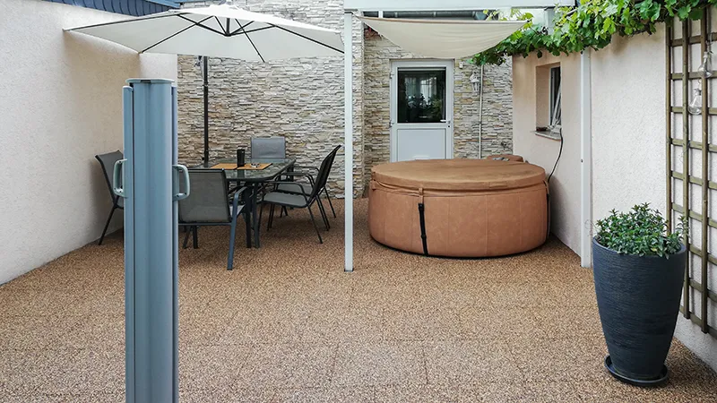 Sous une terrasse couverte, des dalles de terrasse WARCO en granules de caoutchouc liés au PU, déclinées dans des tons beige et brun, créent un sol chaleureux à l’aspect naturel. Un spa gonflable installé transforme l’espace extérieur en un home spa convivial. Les dalles élastiques, antidérapantes et faciles d’entretien absorbent le bruit des pas et protègent du froid. L’ensemble forme un havre accueillant pour se détendre et profiter du plein air.