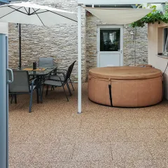 Sous une terrasse couverte, des dalles de terrasse WARCO en granules de caoutchouc liés au PU, déclinées dans des tons beige et brun, créent un sol chaleureux à l’aspect naturel. Un spa gonflable installé transforme l’espace extérieur en un home spa convivial. Les dalles élastiques, antidérapantes et faciles d’entretien absorbent le bruit des pas et protègent du froid. L’ensemble forme un havre accueillant pour se détendre et profiter du plein air.