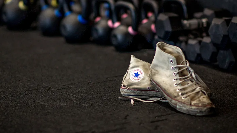 Een paar zwaar versleten Converse ALL STAR sneakers staat op zwarte fitnessvloerbeschermingsmatten van WARCO. Een paar zwaar gebruikte Converse ALL STAR sneakers staat voor een rij halters in de sportschool op zwarte turnmatten van WARCO.