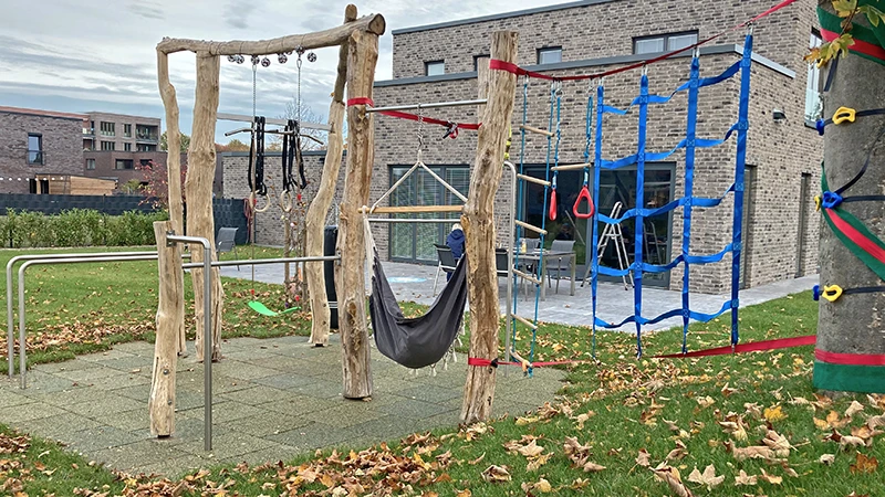 Ein moderner Turn- und Spielplatz bei einem Einfamilienhaus, der mit Gummimatten als Fallschutz von WARCO ausgelegt wurde. Die Matten sind mit einer EPDM-Deckschicht im Farbdesign Savanne versehen, das eine erdige und natürliche Optik vermittelt. Das Spielgerät besteht aus grob bearbeiteten Holzpfosten, an denen verschiedene Kletter- und Schaukelelemente wie Netze, Ringe und eine Hängematte befestigt sind. Die Spielfläche ist von Rasen umgeben und mit herbstlichem Laub bedeckt, während im Hintergrund ein modernes Gebäude mit Klinkerfassade zu sehen ist. Ein auffälliges Detail ist der zu eng gewählte Fallraum um das Spielgerät, der das Sicherheitskonzept des Platzes beeinträchtigen könnte.