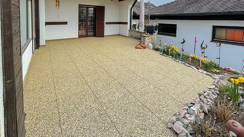 Sur une terrasse partiellement couverte, des dalles de terrasse WARCO en granules de caoutchouc liés au PU affichent une teinte beige chaleureuse évoquant le travertin naturel. Antidérapantes, amortissantes et faciles d’entretien, elles conviennent parfaitement à l’extérieur. Leur structure poreuse favorise l’évacuation rapide de l’eau de pluie, tandis que la surface élastique protège du froid du sol. Sans collage permanent, elles offrent une grande liberté d’aménagement. Le résultat est un espace extérieur confortable à l’ambiance accueillante.