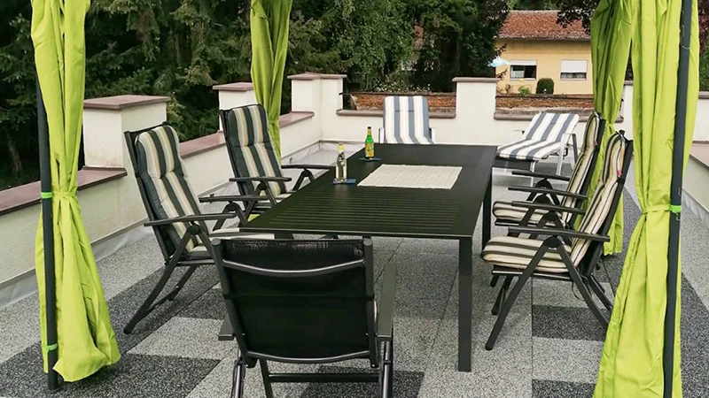 Op een dakterras ligt een vloer van WARCO-terrasplaten in licht- en donkergrijze gemarmerde tinten, vergelijkbaar met graniet. Een baldakijn overspant de zithoek op de elastische, weerbestendige ondergrond. De slipvaste, schokdempende platen kunnen zonder vaste verlijming worden gelegd en dempen loopgeluiden. Zo ontstaat een onderhoudsvriendelijk oppervlak voor ontspannen uren in de buitenlucht.