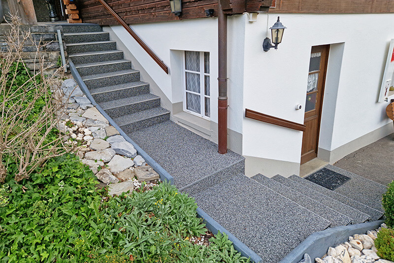 Een grijze trap loopt langs het huis van de kelder naar de eerste verdieping. Planten staan langs de trap.