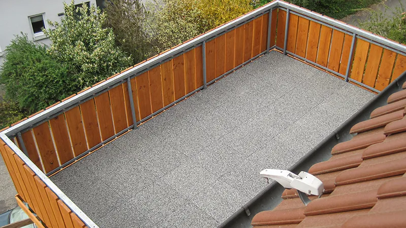 Op de foto is een ruim balkon te zien, dat is voorzien van grijze WARCO-vloerplaten. De omlopende houten bekleding zorgt voor privacy en sluit mooi aan bij de moderne vloer. Dankzij de antislip- en weerbestendige eigenschappen is deze vloer ideaal voor buitengebruik. Bovendien is hij elastisch en vangt hij stappen aangenaam op. Zo ontstaat er een comfortabele en onderhoudsvriendelijke buitenruimte waar men heerlijk kan ontspannen.