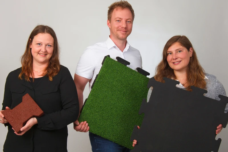 Auf dem Bild sind drei Teammitglieder zu sehen, die verschiedene Bodenbelagsplatten für industrielle Anwendungen präsentieren. Die Platten sind in verschiedenen Farben und Oberflächenstrukturen erhältlich, darunter eine grüne Grasoptik und eine robuste schwarze Platte.