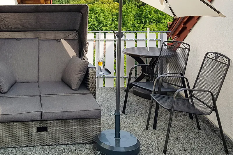Op de foto zie je een balkon met grijze WARCO-vloerplaten. Een comfortabele strandstoel en een klein tafeltje met twee metalen stoelen bieden een plek om te ontspannen en te genieten. De elastische, antislip vloer beschermt tegen vocht en dempt loopgeluiden. Daarnaast is hij onderhoudsvriendelijk en weerbestendig, zodat de buitenruimte er lang uitnodigend uitziet en blijft.
