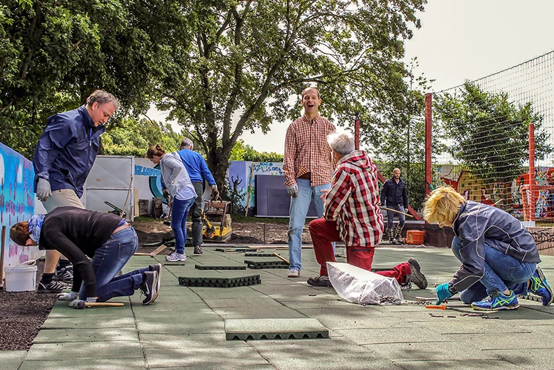 Les parents souriants font eux-mêmes la pose de tapis antichoc WARCO au cour de récréation d’école de leurs enfants.