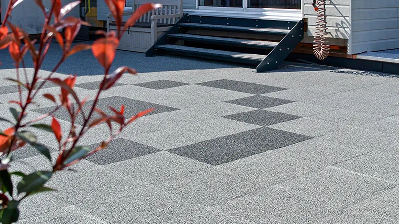 Sur une vaste terrasse, des dalles de terrasse WARCO en granules de caoutchouc liés au PU sont posées en joints croisés, formant un damier saisissant. Les dalles gris clair sont ponctuées de quelques dalles gris foncé dessinant une figure géométrique. Ce revêtement élastique est antidérapant, facile d’entretien et atténue les bruits de pas. De plus, il résiste aux intempéries et convient à un usage toute l’année, apportant davantage de confort en extérieur.