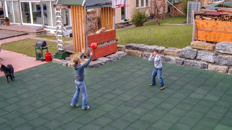 Une femme et un garçon jouent au volley sur un terrain de jeu, fait de dalles vertes de WARCO, dans le jardin privé. Avec les dalles de jeu de ballon WARCO, on peut créer un terrain de jeu polyvalent. Quelle que soit le temps, le terrain est ouvert à tout type d'utilisation sportive.