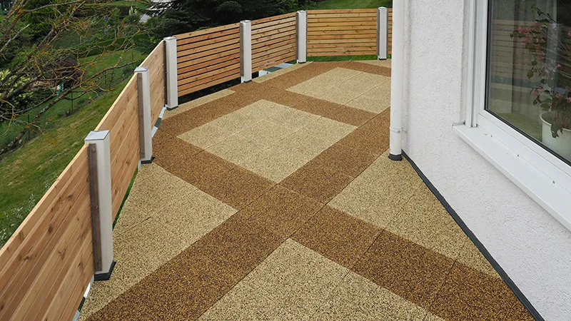 Op de foto zie je een ruim terras met twee kleurvarianten van de WARCO-vloerplaten. Een decoratief patroon in licht- en donkerbruin loopt over het hele oppervlak. De omringende houten balustrade zorgt voor privacy en sluit aan bij de natuurlijke uitstraling van de vloer. Dankzij de antislip- en weerbestendige eigenschappen is deze vloer ideaal voor buitenruimtes en creëert hij een uitnodigende sfeer. Hij is bovendien eenvoudig te reinigen en te leggen. Met weinig onderhoud blijft het een praktische en duurzame keuze.