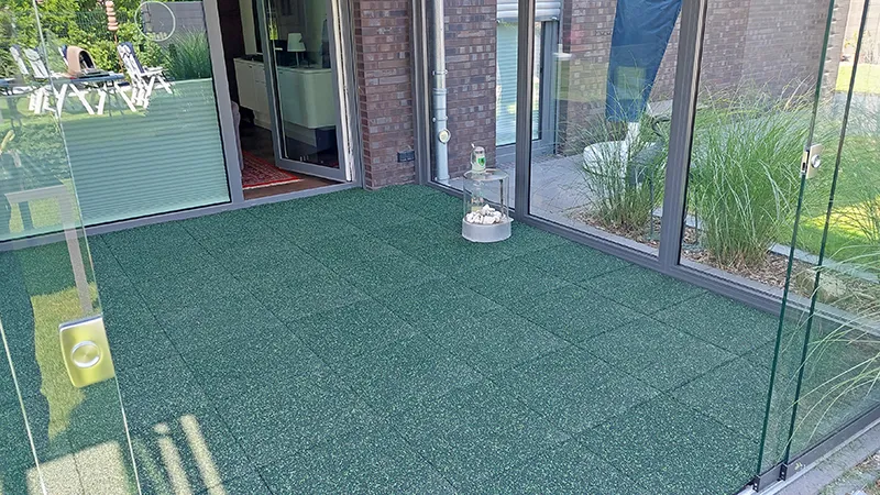 Sur la terrasse se trouve un revêtement en dalles WARCO de couleur verte. Ces dalles maniables sont posées avec un joint croisé classique et maintenues ensemble par un emboîtement caché. Le granulat de caoutchouc offre une surface antidérapante, facile d’entretien et isolante contre le froid du sol, prolongeant ainsi la saison de la terrasse.