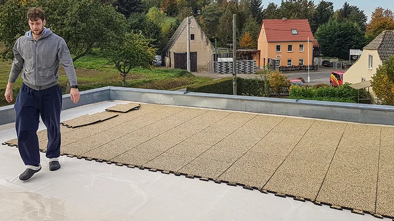 Op deze foto is te zien hoe iemand beige WARCO-puzzeltegels legt op een royaal balkon. Dankzij het nauwkeurig passende puzzelsysteem kunnen de vloerdelen snel en eenvoudig worden samengevoegd. De antislip en weerbestendige toplaag beschermt de ondergrond en biedt een beloopbaar oppervlak voor diverse toepassingen. Zo ontstaat een veilige, comfortabele en onderhoudsvriendelijke buitenruimte, ideaal om te ontspannen. Bovendien zorgt de isolerende werking voor minder loopgeluid.