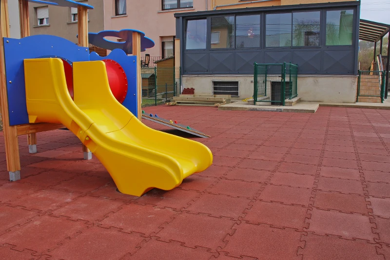 Das Bild zeigt einen Spielplatz mit einem gelben Doppelrutschenturm im Vordergrund, der auf einer Fläche aus WARCO Fallschutzmatten aus Gummi steht. Die Matten sind ziegelrot und durch die Schwalbenschwanzverzahnung an den Plattenrändern formschlüssig zu einem stabilen Plattenteppich verbunden. Im Hintergrund sieht man ein Gebäude. Der Bodenbelag bietet eine stoßdämpfende Oberfläche, die das Verletzungsrisiko bei Stürzen minimiert und gleichzeitig eine langlebige und witterungsbeständige Lösung für den Außenbereich darstellt.