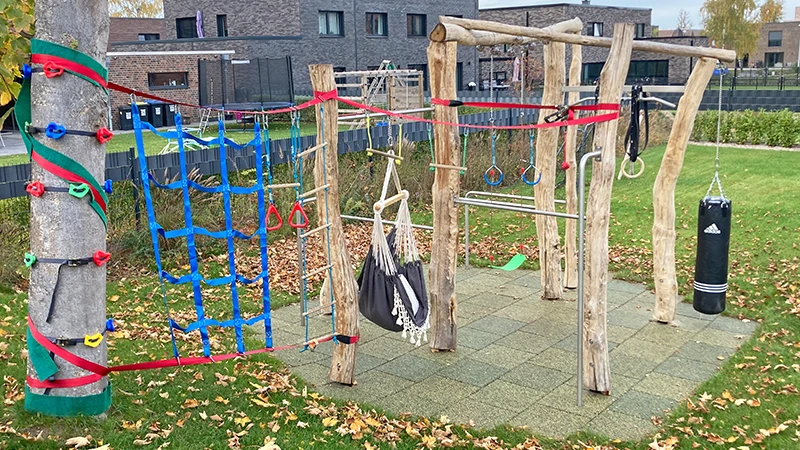 Klimrek met netten ertussen aan een boom. Een bokszak en een schommel en verschillende horizontale stangen hangen aan houten palen.