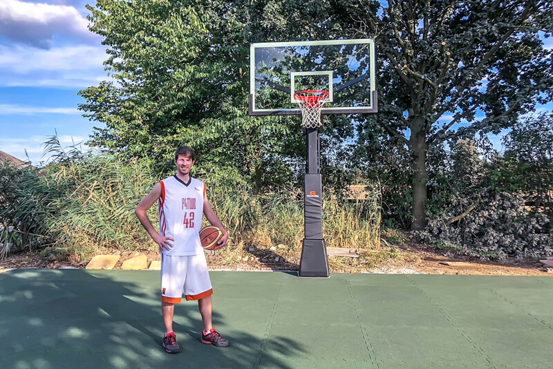 Auf einem professionell mit Ballspielplatten von WARCO gestalteten Basketball-Court im Garten steht ein groß gewachsener Basketball-Spieler mit untergeklemmten Ball.
