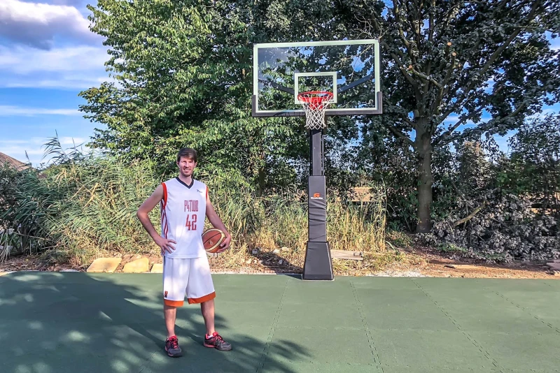 Een lange basketbalspeler met zijn bal eronder geklemd staat op een professioneel ontworpen basketbalveld in de tuin met balpanelen van WARCO.