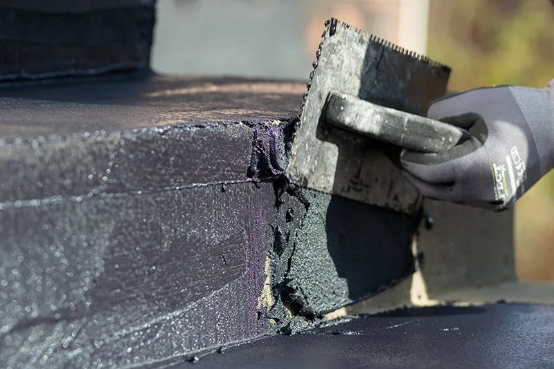 Das Bild zeigt einen Handwerker, der die Gummiabdichtung ALLESDICHT von WARCO mit einer Zahnkelle auf eine Betonfläche aufträgt. Die schwarze, elastische Abdichtungsmasse wird in einer gleichmäßigen Schicht aufgetragen, um eine wasserundurchlässige und hochreißfeste Schutzschicht zu bilden. Die Struktur der Abdichtungsmasse ist deutlich zu erkennen, während die Zahnkelle dabei hilft, das Material gleichmäßig zu verteilen und Haftung zu gewährleisten.