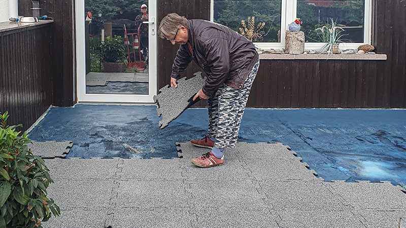 Une terrasse est en cours de rénovation. Une dame âgée pose la prochaine dalle de terrasse de 18 mm d'épaisseur avec emboîtement sur la terrasse déjà partiellement recouverte de nouvelles dalles. Une feuille d'étanchéité sert de support pour le nouveau revêtement de sol en dalles de caoutchouc. L'emboîtement précis des dalles permet une pose facile et stable.