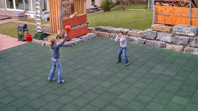 Une femme et un garçon jouent au volley-ball sur un terrain de sport fait de dalles vertes de jeu de ballon de WARCO dans leur jardin privé. Quel que soit le temps, le terrain est ouvert à toutes sortes d'activités sportives.