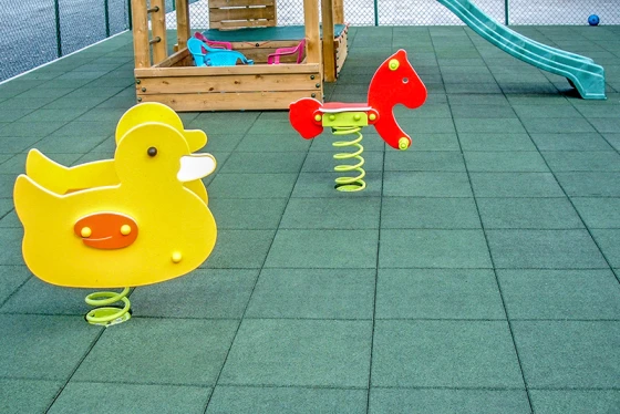 Das Bild zeigt einen Spielplatz, dessen Boden mit grasgrünen WARCO Fallschutz-Gummimatten ausgelegt ist. Im Vordergrund sind zwei bunte Wipptiere auf Federn zu sehen: eine gelbe Ente und ein rotes Pferd. Im Hintergrund befindet sich eine kleine Holzstruktur mit bunten Sitzgelegenheiten und einer grünen Rutsche. Der gesamte Spielplatz ist von einem Maschendrahtzaun umgeben, der für Sicherheit und Schutz sorgt. Die Fallschutzmatten bieten eine stoßdämpfende Oberfläche, die die Kinder vor Verletzungen schützt und gleichzeitig für ein attraktives grünes Erscheinungsbild des Spielplatzes sorgt.