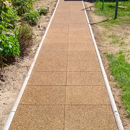 Ein gerader Gartenweg aus quadratischen Gummiplatten mit seitlicher Bordsteineinfassung führt durch das Beet. Die braunen Platten aus Gummigranulat bilden eine pflegeleichte, wetterbeständige und rutschhemmende Oberfläche. Der Weg sorgt für sauberes, sicheres Begehen bei jedem Wetter und grenzt die bepflanzten Flächen klar von der Rasenfläche ab. Ideal für Gärten, Höfe und Zugänge.