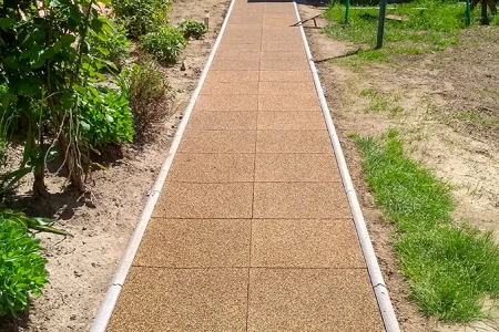 Een recht tuinpad van vierkante rubbertegels met zijafwerking van boordstenen loopt door het perk. De bruine tegels van rubbergranulaat vormen een onderhoudsarme, weerbestendige en antislip ondergrond. Het pad zorgt voor schoon en veilig lopen bij elk weer en scheidt de beplante perken duidelijk van het gazon. Ideaal voor tuinen, erven en toegangen.