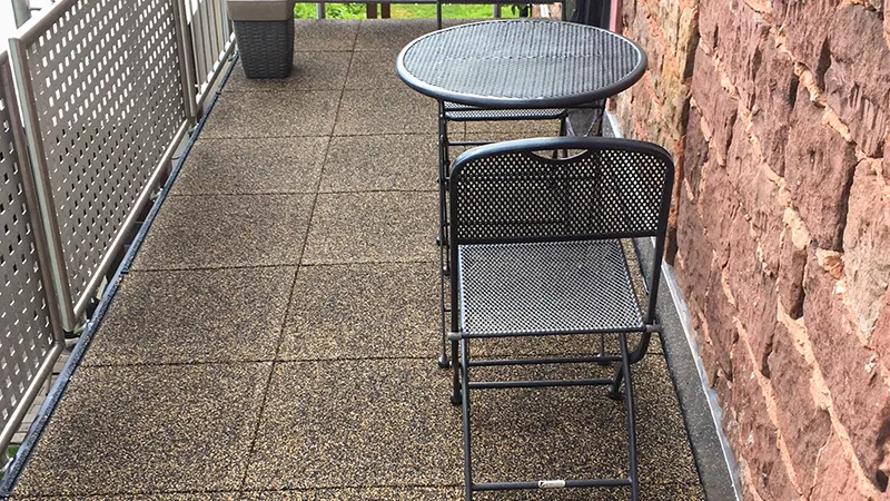Op de foto zie je een smal balkon dat is voorzien van bruin-beige WARCO-tegels. Een kleine, opklapbare metalen tafel met bijpassende stoel biedt een compacte zitplek. De stevige metalen balustrade en de natuurstenen muur vormen een harmonieus geheel. Dankzij de antislip- en weerbestendige eigenschappen is deze vloer ideaal voor buitenruimtes, en hij is eenvoudig te reinigen en te plaatsen.