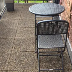 Op de foto zie je een smal balkon dat is voorzien van bruin-beige WARCO-tegels. Een kleine, opklapbare metalen tafel met bijpassende stoel biedt een compacte zitplek. De stevige metalen balustrade en de natuurstenen muur vormen een harmonieus geheel. Dankzij de antislip- en weerbestendige eigenschappen is deze vloer ideaal voor buitenruimtes, en hij is eenvoudig te reinigen en te plaatsen.
