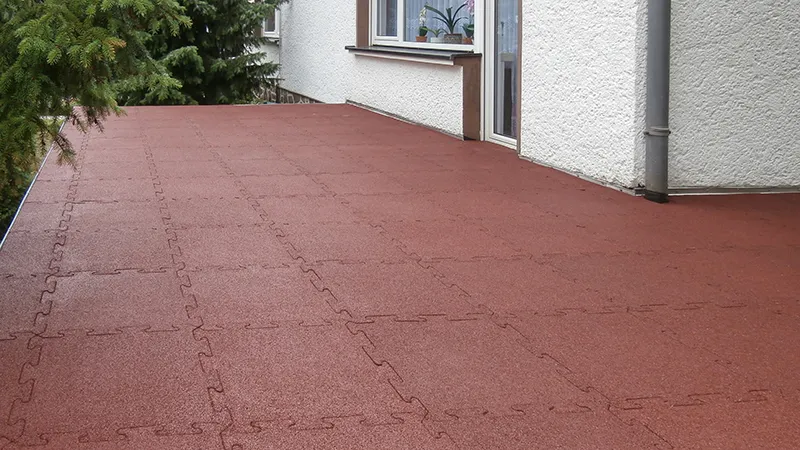 Sur une terrasse de la maison, des dalles de terrasse WARCO en granules de caoutchouc liés au PU, dans une teinte rouge brique classique, sont posées. Grâce à l’emboîtement puzzle, les dalles restent solidement en place. Le revêtement élastique est antidérapant, amortissant et résistant aux intempéries. Les dalles peuvent être facilement découpées sur mesure et posées sans adhésif permanent.