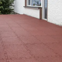 Sur une terrasse de la maison, des dalles de terrasse WARCO en granules de caoutchouc liés au PU, dans une teinte rouge brique classique, sont posées. Grâce à l’emboîtement puzzle, les dalles restent solidement en place. Le revêtement élastique est antidérapant, amortissant et résistant aux intempéries. Les dalles peuvent être facilement découpées sur mesure et posées sans adhésif permanent.