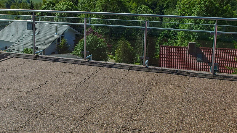 Deze foto toont een royaal balkon met een transparante balustrade, vanwaar je uitkijkt op omliggende daken en groen. De vloer is bekleed met WARCO puzzeltegels van rubbergranulaat in een warme bruintint. Dankzij de in elkaar grijpende randen ontstaat er een naadloos oppervlak dat zowel antislip als weerbestendig is—ideaal voor buitenruimtes. Bovendien biedt de vloer schokdemping en goede geluidsisolatie. Door het onderhoudsvriendelijke oppervlak is het een duurzame en comfortabele keuze voor balkons en terrassen.