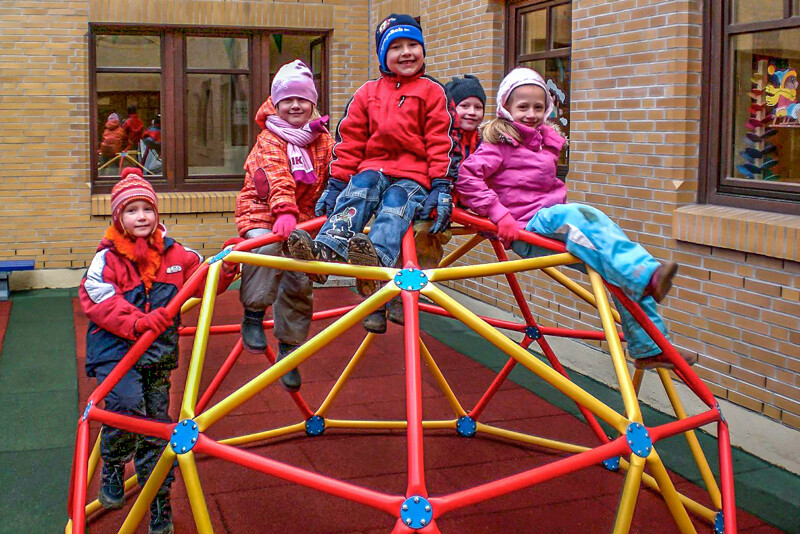 Im Innenhof einer Kindertagesstätte steht ein kugelförmiges Klettergerüst aus Rohren auf roten und grünen Fallschutzplatten. Eine Gruppe von Kindern auf dem Klettergerüst lächelt den Betrachter an.